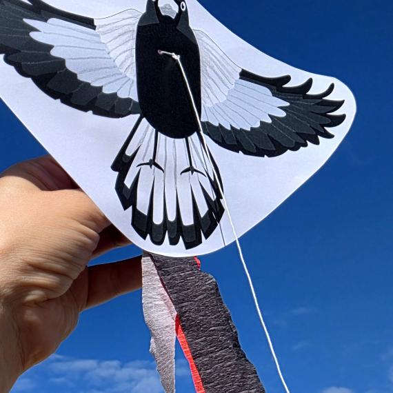 A hand holding up a kulbardi/magpie kite craft activity with a blue sky background, available at Fremantle WA Day celebrations in 2026. 