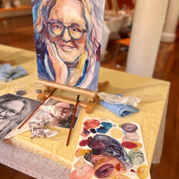 A painting of a woman's face sits on an easel on the table. The woman has grey hair and a blue shirt on, she is smiling and propping up her chin with her hand. Around the painting on the table is a pallette filled with paints and a selection of brushes 