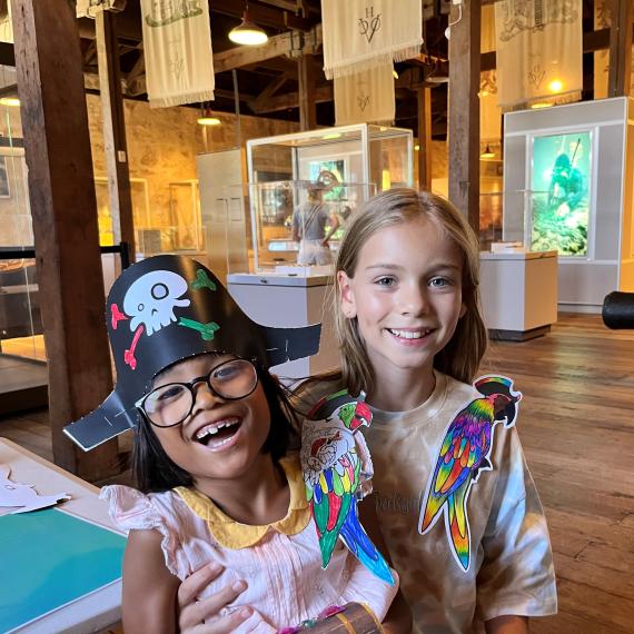 Two girls stand together smiling, wearing paper parrots on their shoulders. One is wearing a pirate hat too.  Behind them are display cases, a lightbox with a diver and historical flags are hung from the roof. The floor is wooden and the walls made of limestone. 