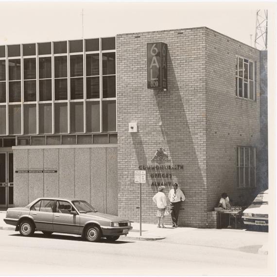 a black and white photograph of a building. the building has a sign on it 6AL and another sign Commonwealth Offices Albany. There are cars partked outside the building on the street and a few people standing next to the building
