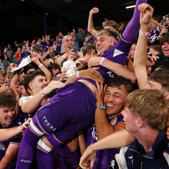 Perth Glory player celebrates with supporters in the stands after a match, surrounded by a cheering crowd in club colours.